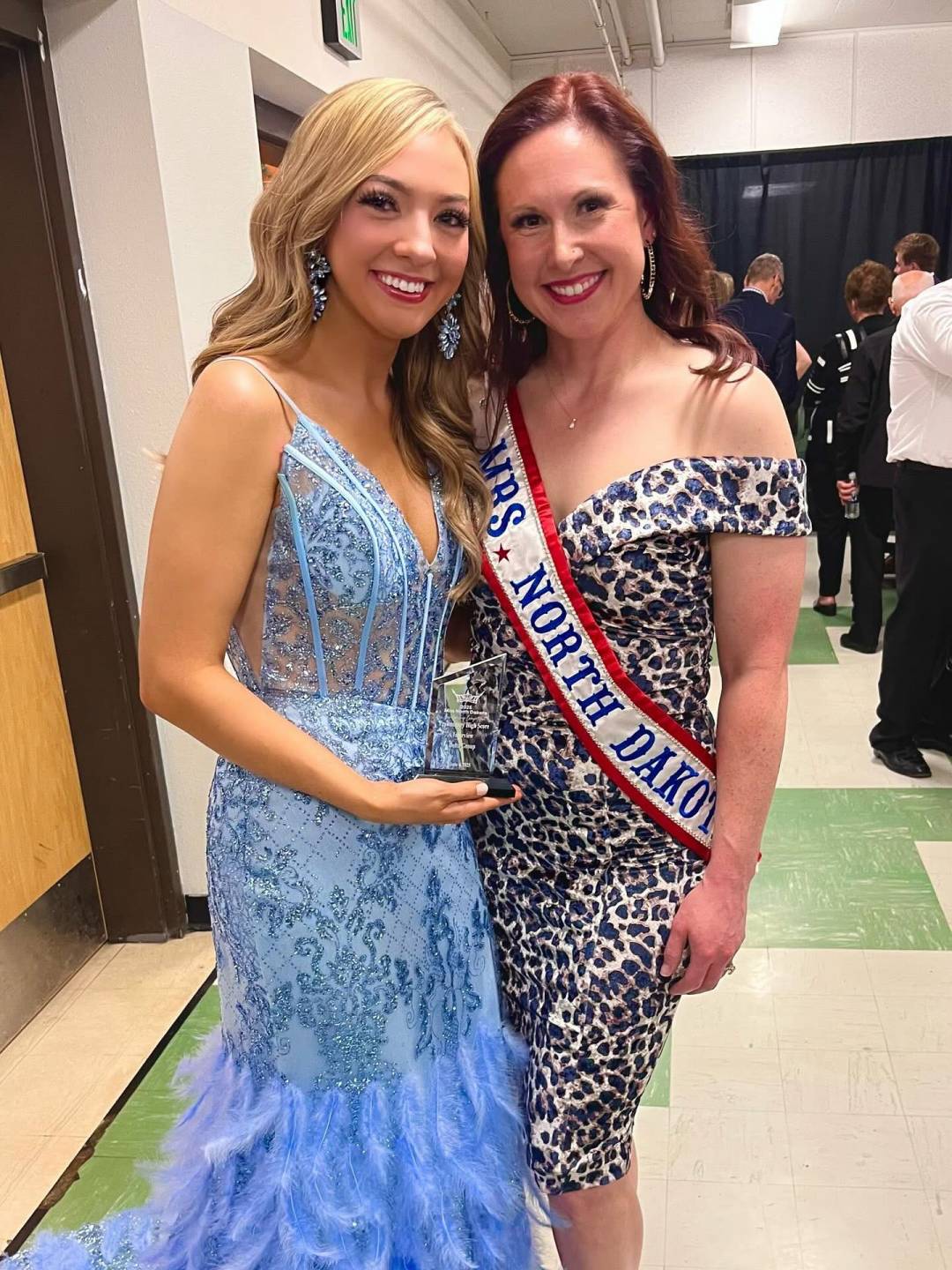 Dana Henry, wearing a leopard print off-the-shoulder dress, is crowned Mrs. North Dakota at the state pageant by the previous titleholder, who is dressed in a light blue sequined gown.