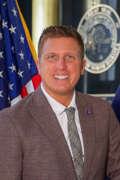 Caucasian male with dark blond hair and blue eyes, smiling. He is wearing a white collared shirt, a light tan suit jacket, and a tie. An American flag is visible in the background.