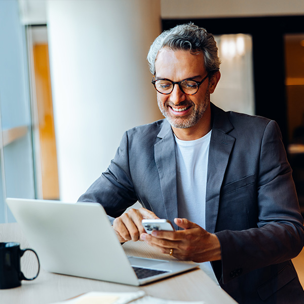A happy man wearing glasses and a charcoal blazer checks his smartphone while sitting next to his laptop at a coffee shop.